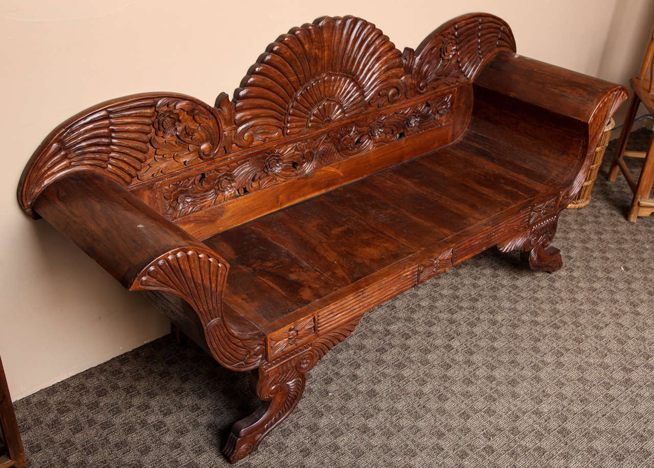 A 19th century settee from Jakarta, Indonesia, where it was carved out of teak wood. This settee's shape is a traditional Indonesian one, displaying scroll arms and wing-like patterns on its back. The piece is supported by carved legs adorned with