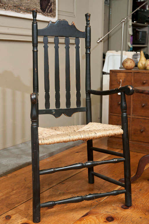 An early 18th c. old grungy surface black painted New England bannister back armchair. The double-arched crest above four shaped vertical balusters flanked by turned stiles with finials. This beautiful chair retains an old crusty black paint surface