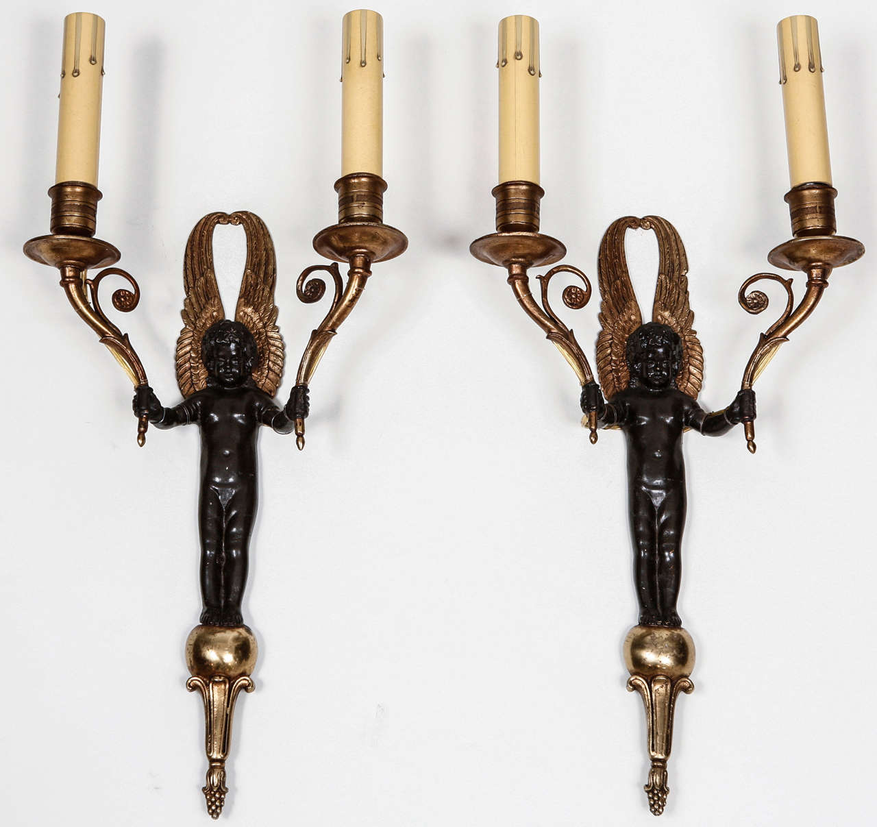 An interesting pair of figural wall sconces with each having two candle holders that have been french wired for lights.  The figures hold a light in each hand, having their wings raised as they stand on an orb. These sconces will add to any setting
