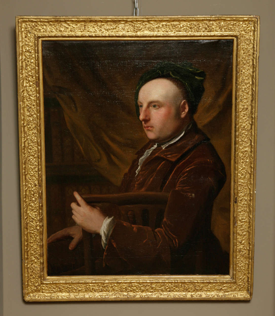 Circle of Thomas Gibson (British, c.1680-1751). Sensitively observed and painted portrait of an artist, seated sideways on a mahogany chair in a library, half length, in a rust velvet coat and green velvet cap. Oil on canvas, bears remains of 19th
