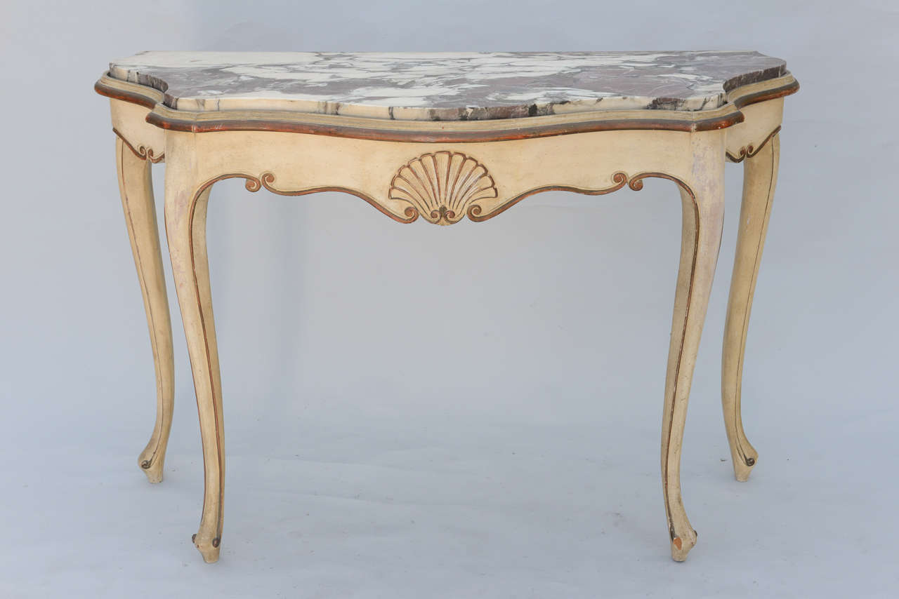 Pair of console tables, distressed painted finish and parcel gilt, each having freeform top inset with thick rouge marble, apron centered with carved scallop shell, flanked by serpentine scrolls, raised on cabriole legs.