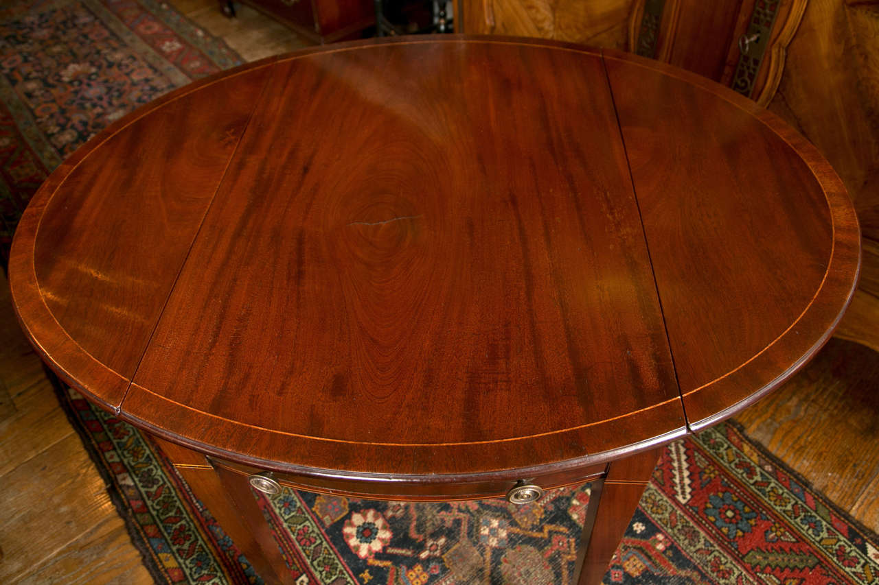 A narrow boxwood strand separates the mahogany self-band from the center field of this delicate Pembroke table. With its shaped-front drawer following the curvature of the top and its narrow, tapered legs, this table has a cohesive balance that