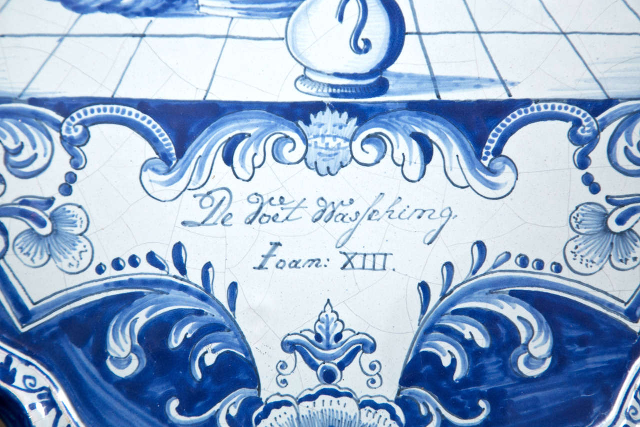 This large blue and white delft plaque is in a cartouche  shape.  It depicts Jesus washing  the feet of possibly a  diciple as others look on. The legend is written in  Dutch.