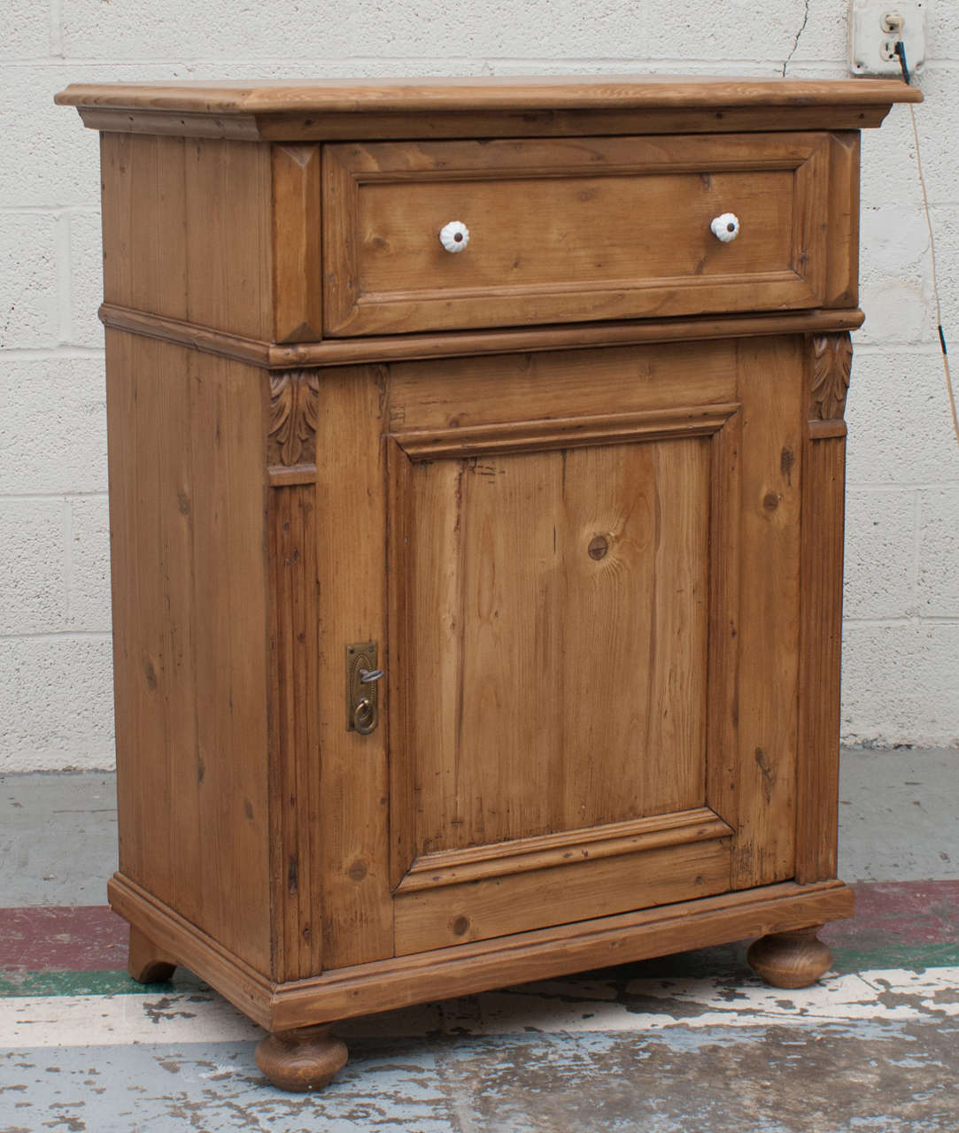 A small pine storage cupboard featuring a single hand-cut dovetailed drawer with original ceramic knobs above a single panelled door flanked by fluted moulding and acanthus leaf corbels.  The feet can be removed to adjust the height for use as a