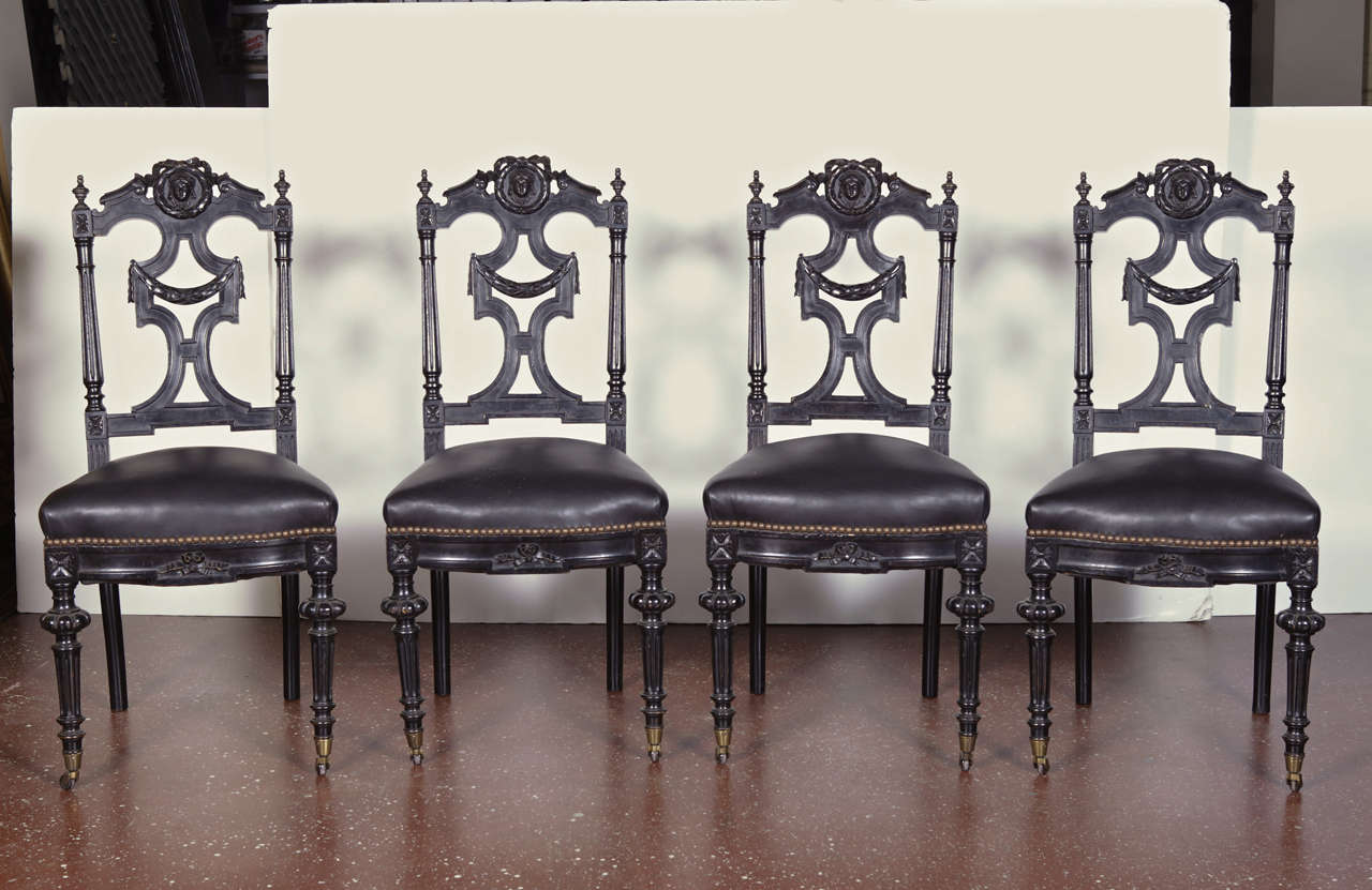 PAIR of 19th c. ebonized side chairs, ornately carved with ribbon and garland motif, masks and finials. Turned front legs with castors. Upholstered in black leather with nail-head trim. Circa 1860.