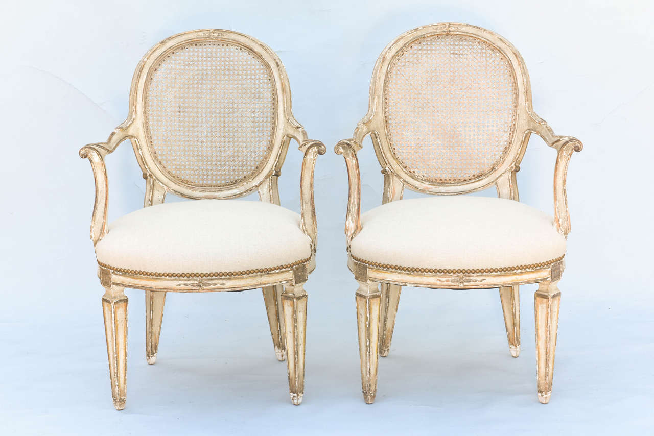 Pair of open armchairs, distressed painted finish, with parcel gilt, each having a fielded frame surrounding oval caned back, outswept carved arms, on scrolling terminals, stuff-over seat upholstered in linen, raised on tapering square