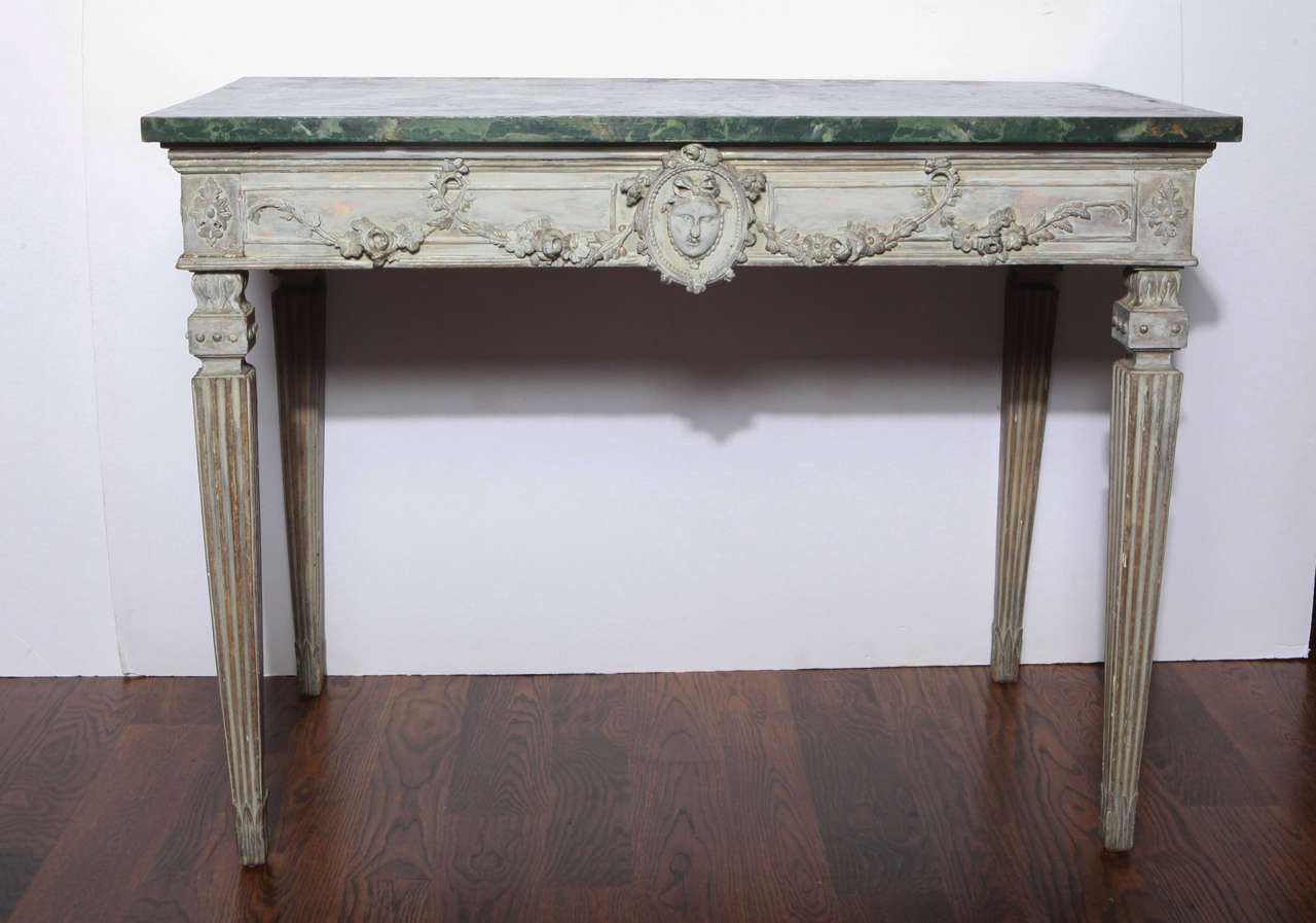 Painted wood console with green marbleized top.  Carved wood apron with draped garland and head at center.  Fluted tapered legs.