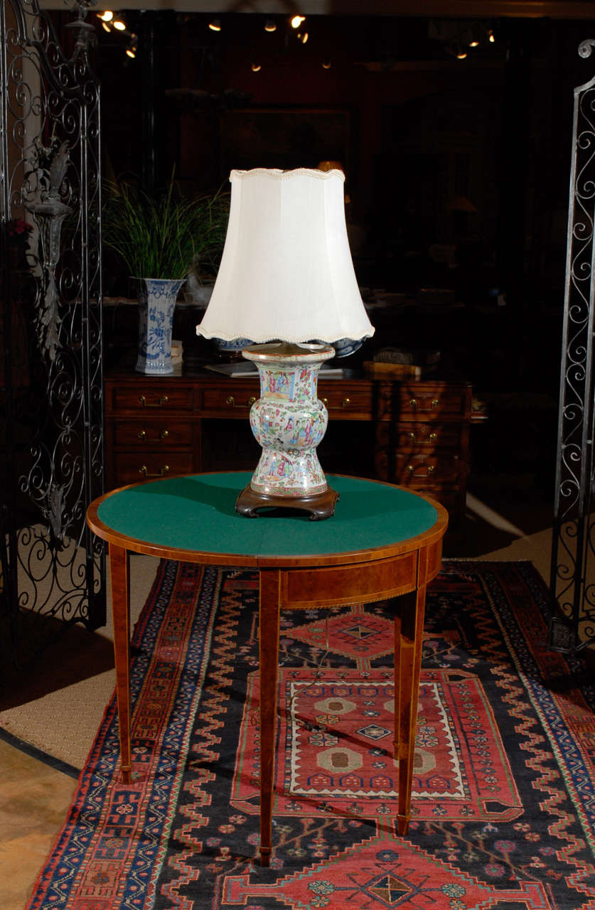 A beautiful large porcelain 19th century rose Mandarin lamp sitting on a wooden Chinese brown stand. The lamp is decorated with people and flowers in primarily shades of green, pink and blue with a cream background. The silk shade is new.
