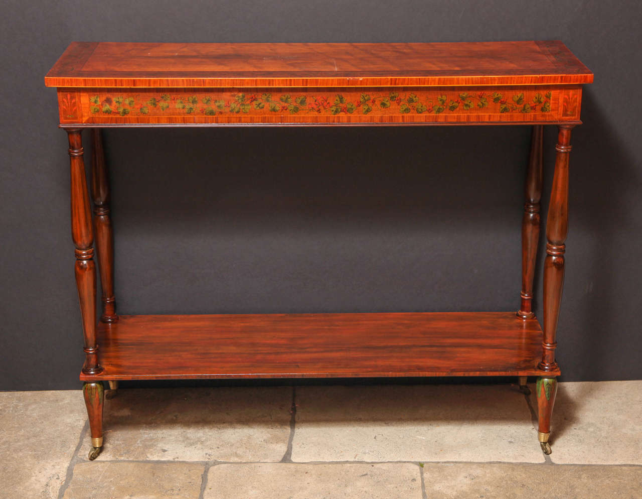 A pair of Adam style painted and crossbanded satinwood console or side tables with scolling foliate decorated top and frieze on turned supports with a shelf stretcher and tapering feet ending in casters.