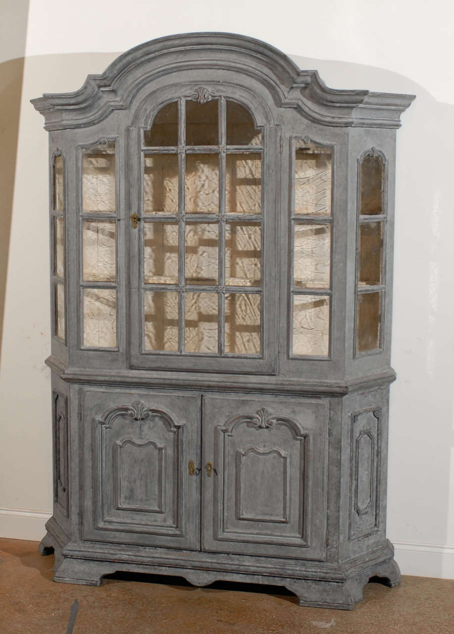 A Swedish Rococo style painted vitrine cabinet with original glass and lower wooden doors from the late 19th century. This Swedish cabinet is topped with an exquisite bonnet-shaped pediment, surmounting a glass door cabinet with canted sides. The