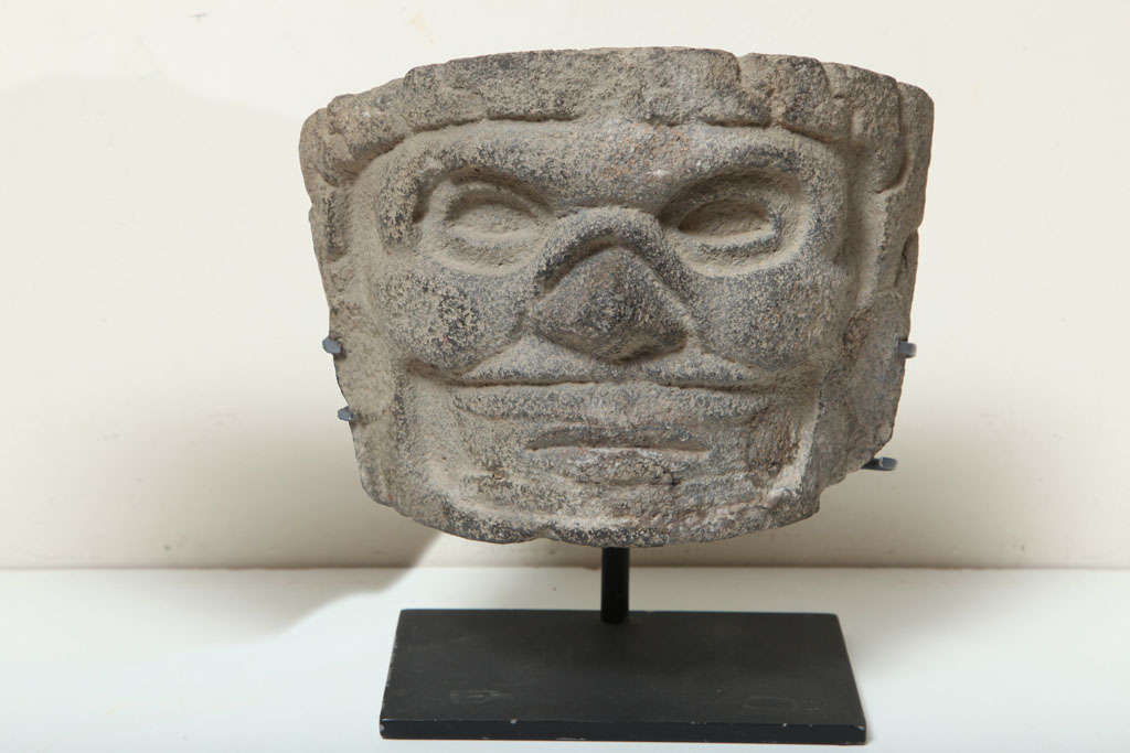 Carved from gray volcanic stone is a central rounded face, wearing a flayed skin mask over face and ear discs. 

Ex Private New York Collection.
 A rare and expressive example. 

Ex Alan Long Collection NYC