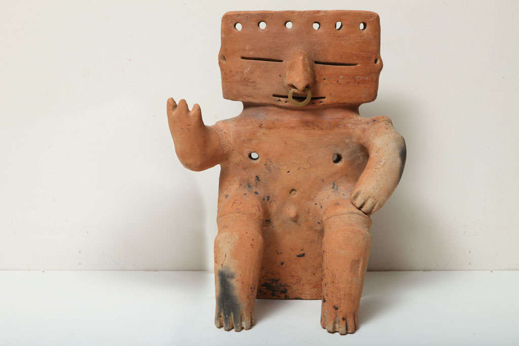 Abstract slab figure, seated with cylindrical protruding limbs and body piercing. He holds right hand in upward gesture. His legs, at knees are constricted. 

Ancient gold nose ring through pierced septum. 


 A handsome example. 

Compare