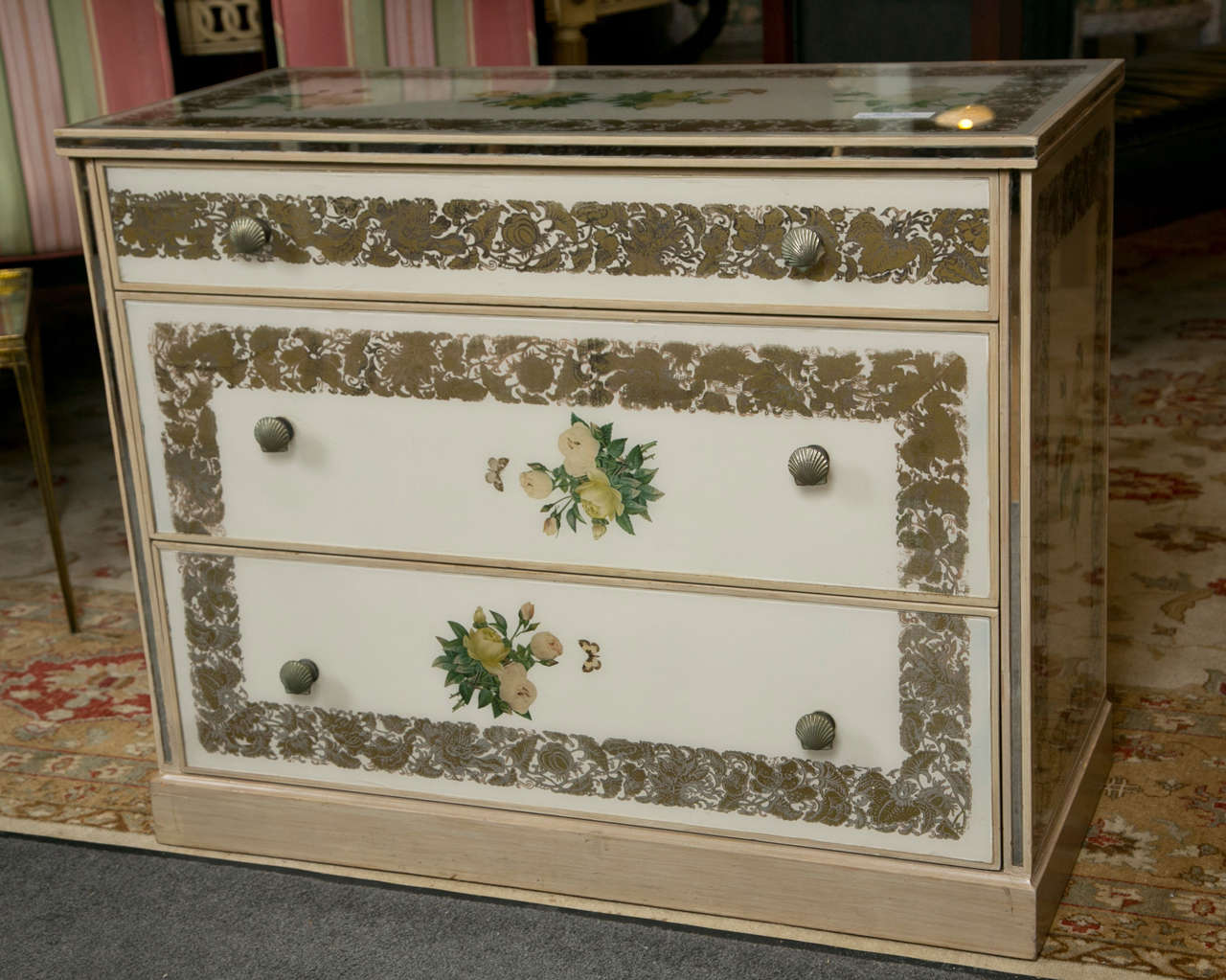A beautiful polychromed chest of drawers in the Art Deco style, decorated with reverse-paint mirror veneers, the conforming case fitted with three drawers each decorated with shell-form pulls, raised on block base.