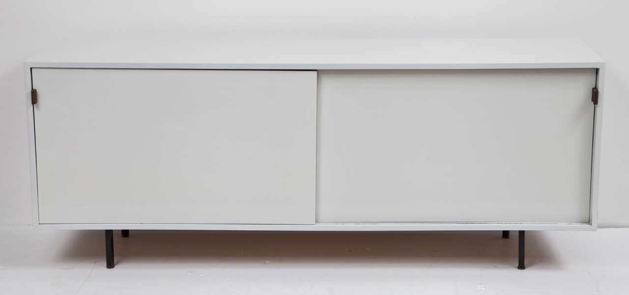 All white credenza with two sliding doors and black metal legs by Knoll.  USA, circa 1960.  Signed.  Features white semi-gloss lacquer finish with original brown leather door handles.  Interior adjustable shelves and drawer.