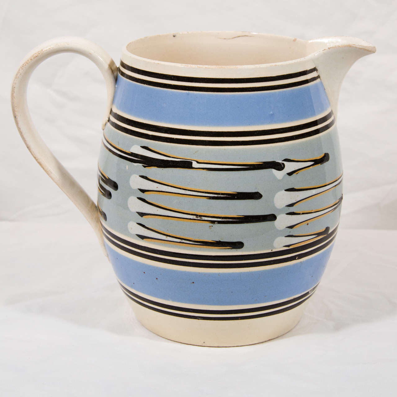 A slip covered, machine turned Mocha jug with broad bands of light blue and thin lines of black at the top and bottom. The center with slip cup decoration in white, black and orange on a light blue gray ground.
For an almost identical jug with