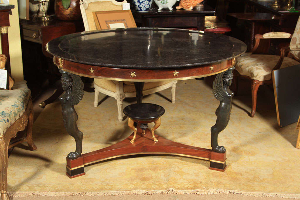Table de centre Empire du début du XIXe siècle en forme de guéridon, pieds monopodes de forme féminine patinés, plateau en marbre, placage d'acajou et décor appliqué en métal doré.