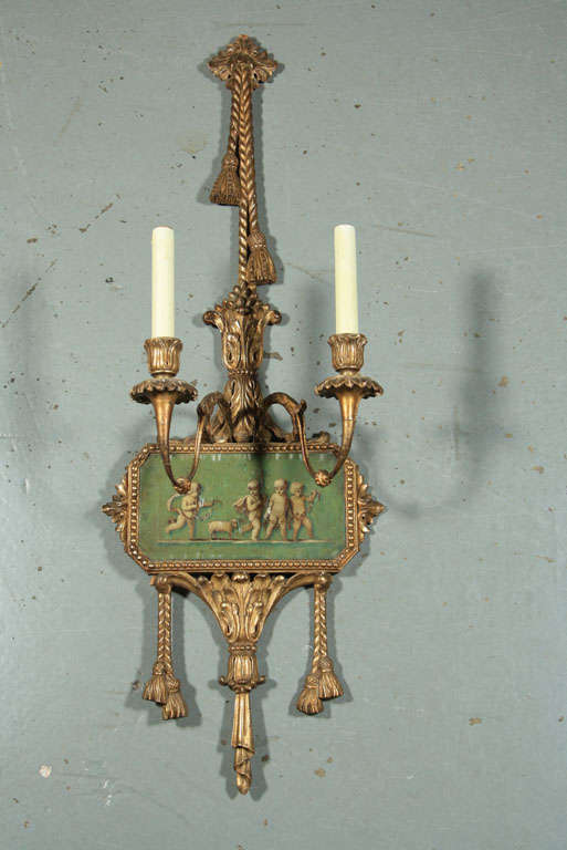 A pair of Adams style, rope and tassel, carved and gilt two arm wall sconces with wooden panels painted with grisaille figures of cherubs on a green backgroud.