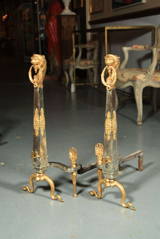 A striking pair of French silvered steel and bronze dore Empire andirons with lions head finials mounted on columns set on snake form feet.