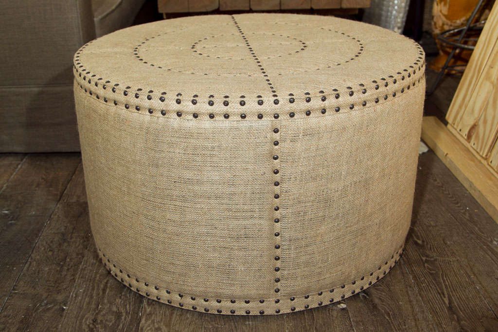 Round ottoman upholstered in burlap with decorative nailhead trim.