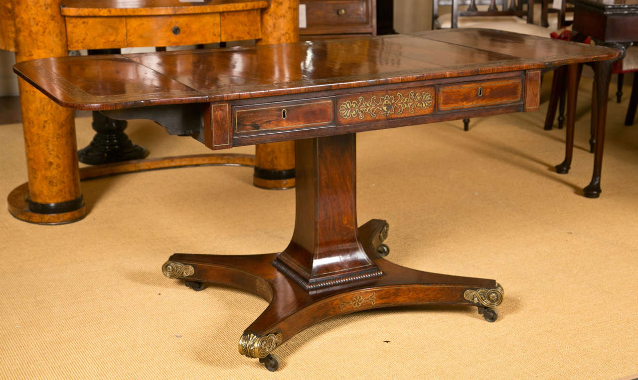 An English Regency period rosewood and brass inlaid sofa table on platform base ending in brass scrolled feet, the stem with brass line inlay and all drawers also brass inlaid, circa 1830. When closed 36 inches wide and 60 inches when open.