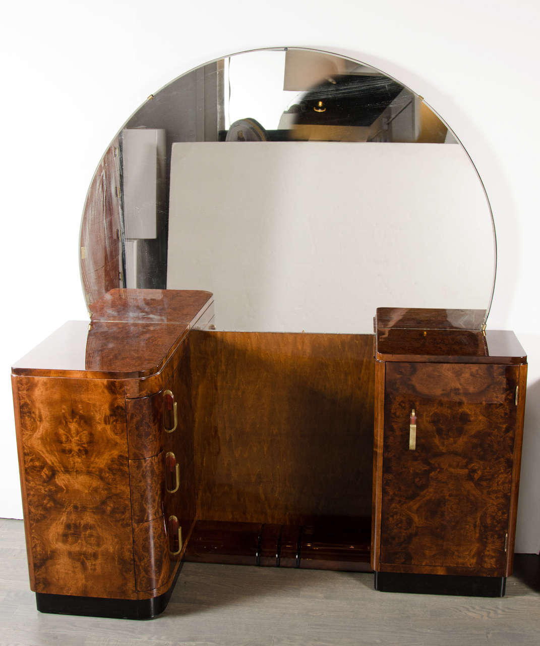 This  Art Deco Streamlined Vanity in the manner of Donald Deskey features a large rear mounted circular mirror, a seating nook with foot rest with stylized Art Deco detailing and a cabinet opposite a set of three drawers. It has brass and bakelite