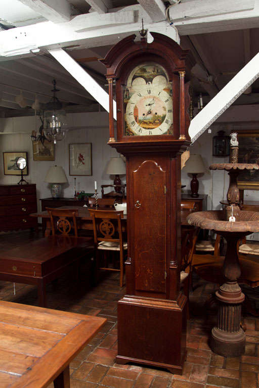 This English, oak tall case clock has an elaborately painted face with both sides of the globe above a circular dial. The bonnet has a broken arch supported by columns that flank the face and are repeated as quarter columns on the case, which itself