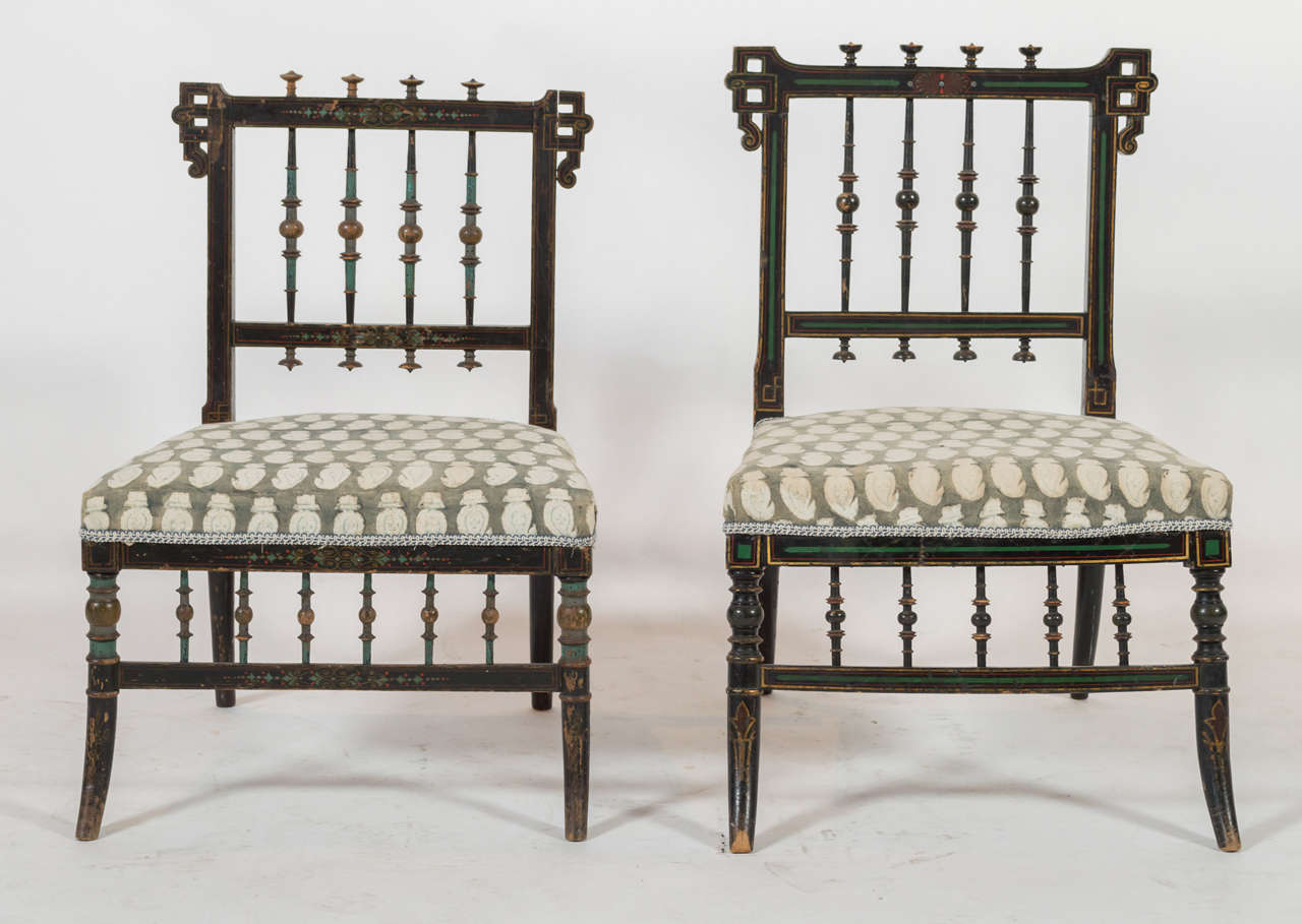 A pair of two small parlor chairs with patterned upholstery. Both chairs are similar, but one is slightly smaller than the other. Seat upholstery is bordered with a blue and white embroidered lace. Wood frame is decorated with faded painted designs.