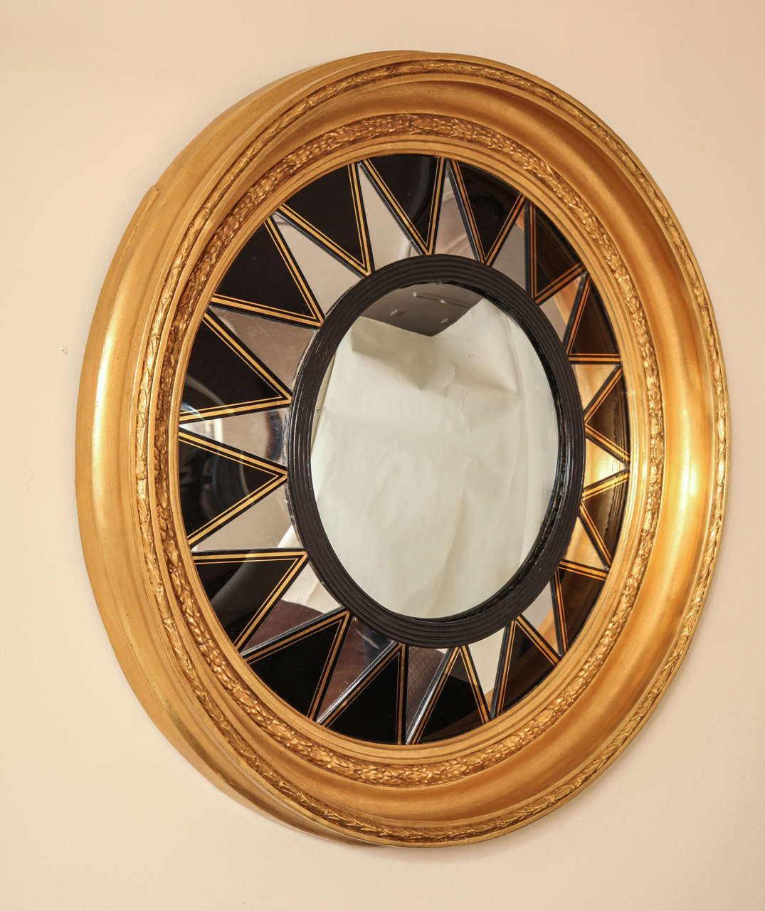 An English Regency period convex mirror, the round center glass framed by ebonized reeded molding surrounded by triangular black plexi elements radiating from center and having gilt stripe details. The outer frame with carved wood and gesso leaf tip