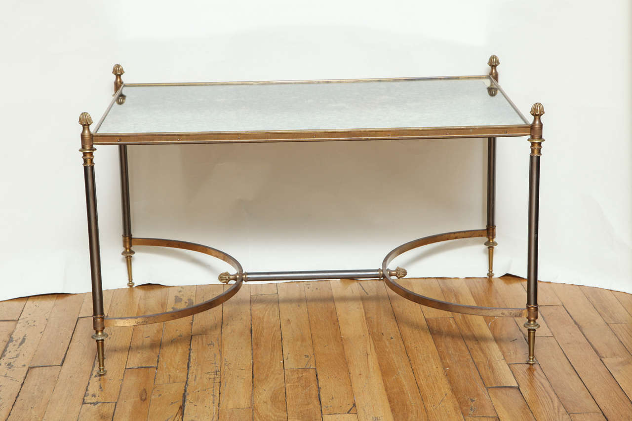 A French Louis XVI style coffee table with antique smokey mirror top having gilt bronze edge supported by steel legs having gilt bronze finials and turnings. The legs supported on the lower section by two c-shaped elements in gilt bronze joined by