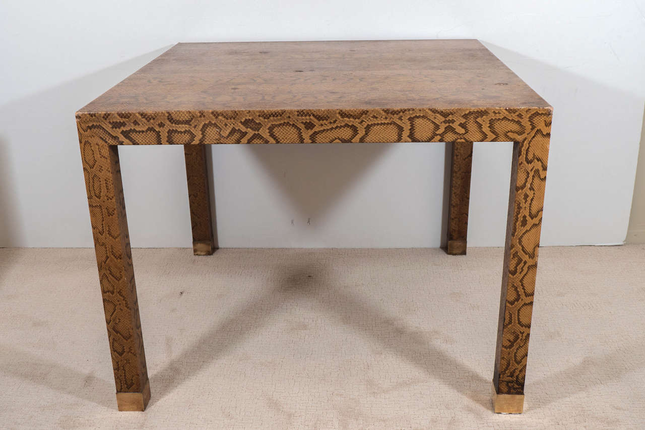 A vintage game table, produced circa 1970's, inspired by designer Karl Springer, upholstered in snakeskin on brass-plated feet; the simplicity of the modern geometric shape of the table and legs creates a unique contrast with the natural patterns of