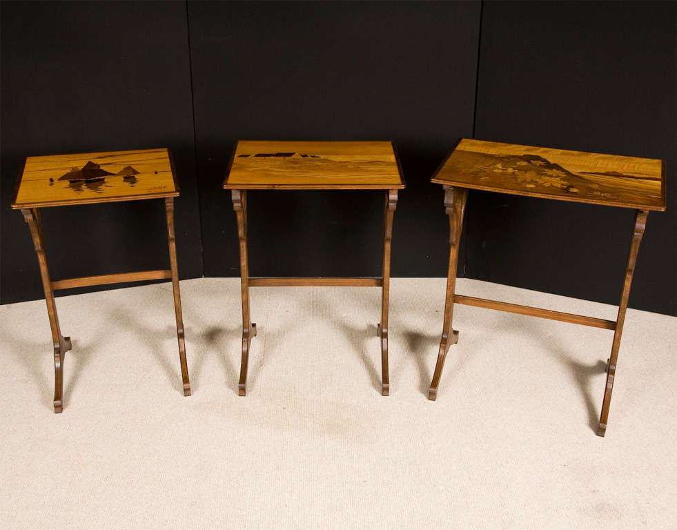 A nest of three Marquetry Occasional Tables by Emile Galle (1846-1904). Each graduated table with rectangular top. The marquetry inlaid in specimen woods with a design of; holly in a mountain landscape, cottages in a landscape, and boats at