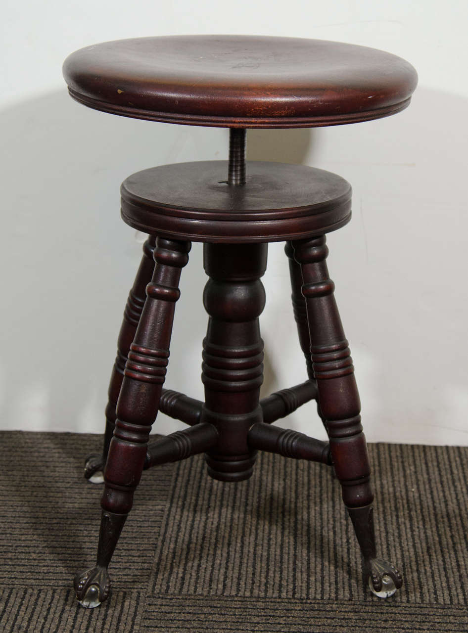 An antique adjustable piano stool in mahogany turned wood embellished with claw and ball feet, and detailed with a bird talon holding a clear glass marble or ball. Legs and base are decorated with carved rings. 

In good condition with age