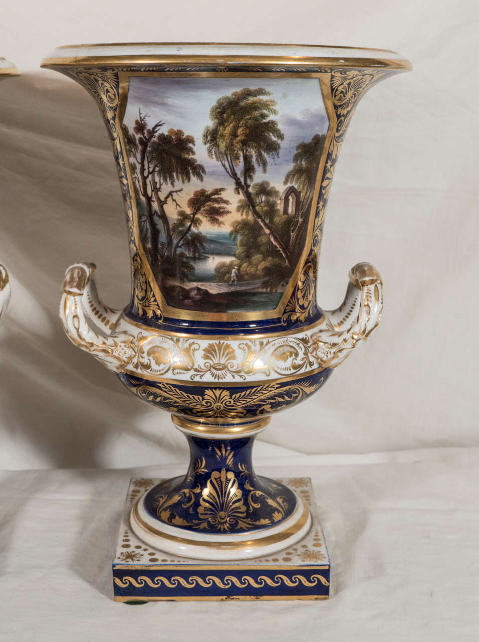 Each urn with square base, scroll handles, a deep blue and gilt ground, and a beautifully painted landscape scene. The scenes 