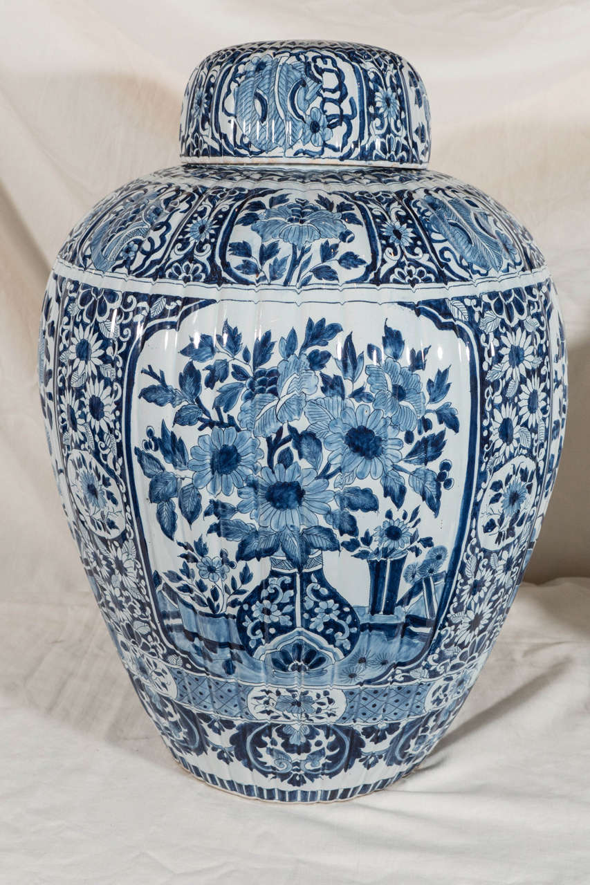 A pair of Dutch delft covered ginger jars ribbed and painted in dark cobalt blue. Decorated with panels showing a traditional image of a flower filled vase placed on the garden terrace. Separating the panels are bands of deep blue flowers and