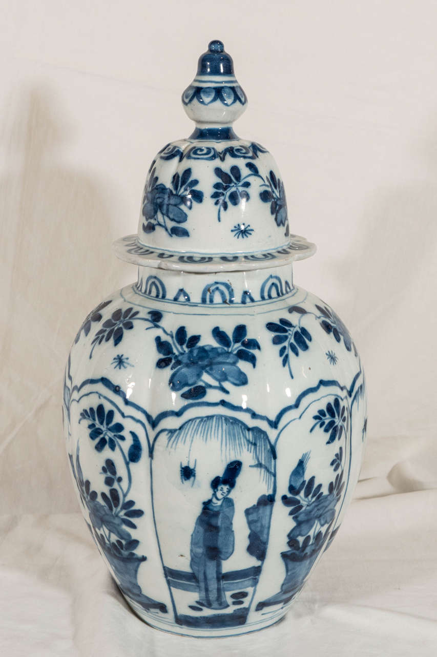 A covered vase decorated in cobalt blue with lobed panels alternately showing a Long Eliza in a garden setting and a flower filled vase. The shoulder and cover are painted with flowers on the vine. The cover topped with a traditional knop