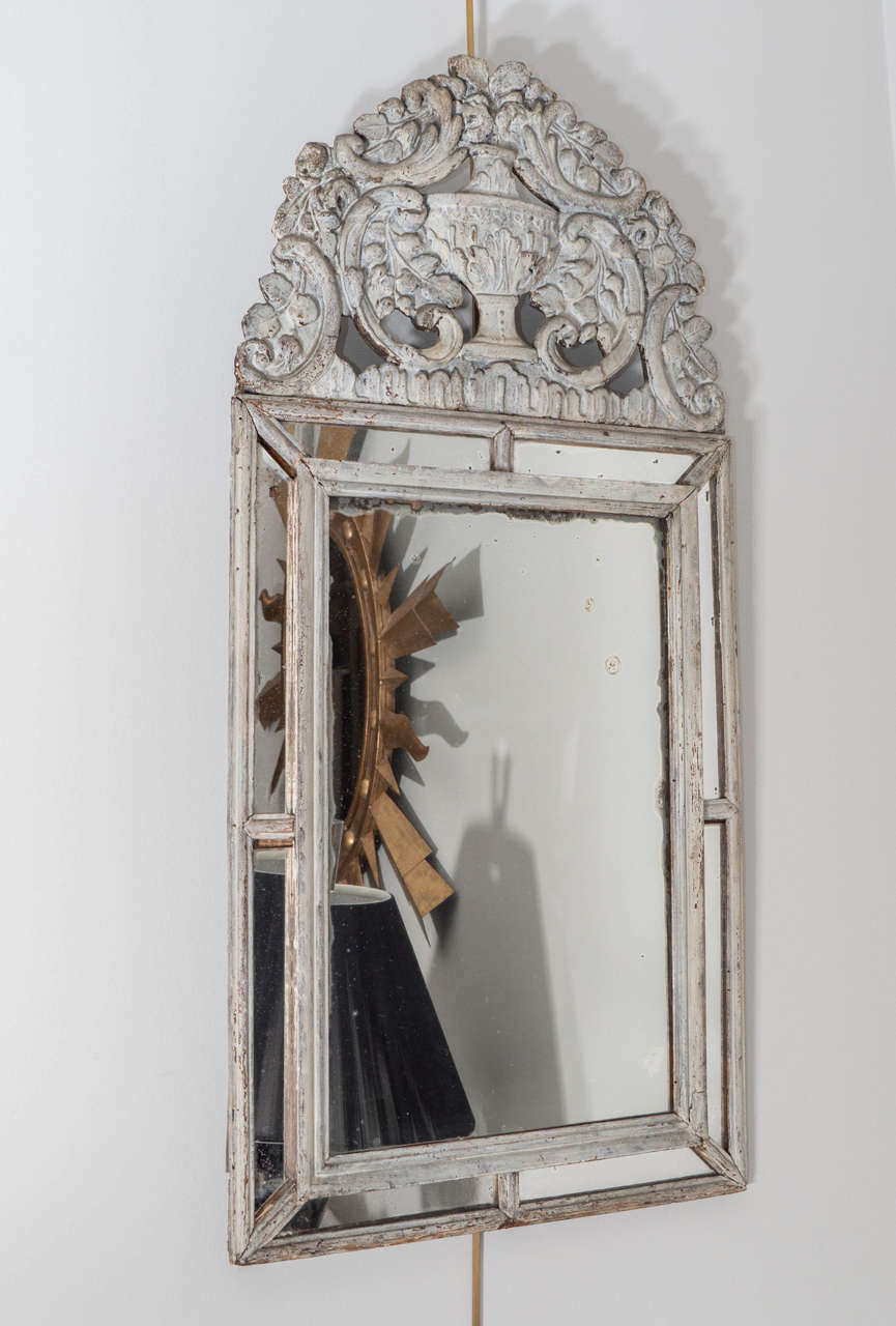 A lovely carved Swedish mirror, painted in a soft gray/white. The glass has a wood frame and is surmounted by a carved urn and a floral motif.