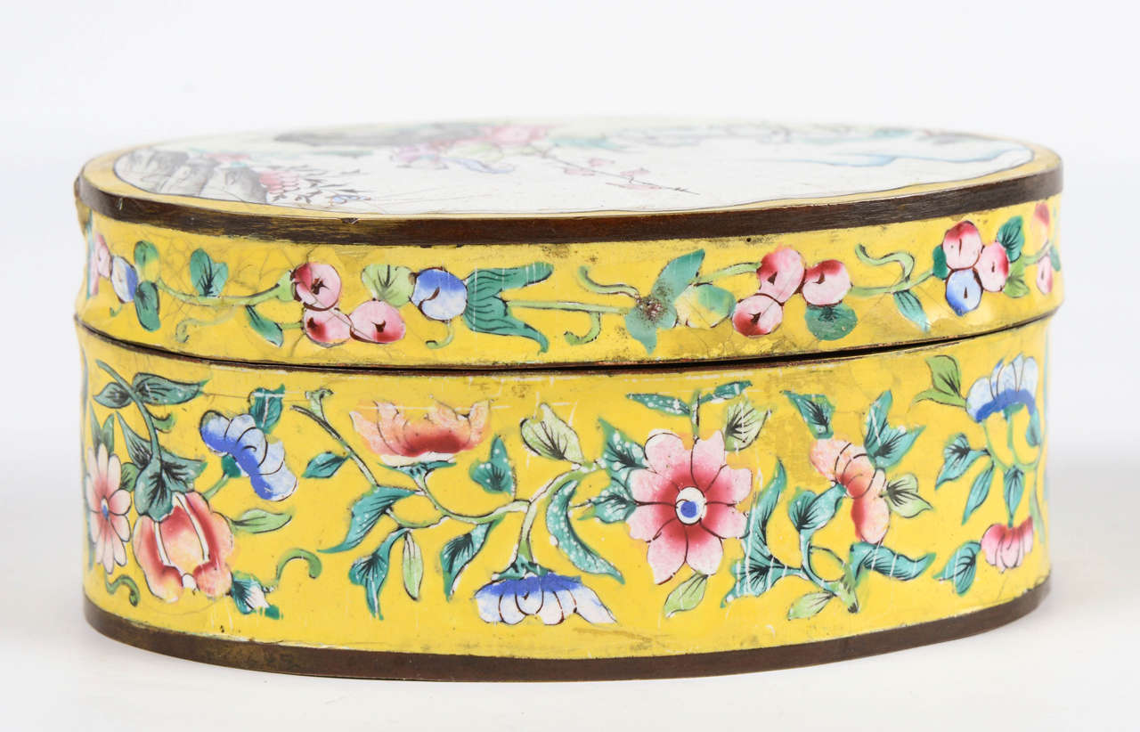 Oval box in canton enamel with a yellow background and polychrome floral decor. The cover has a lively landscape of people and animals. The interior is in blue enamel.