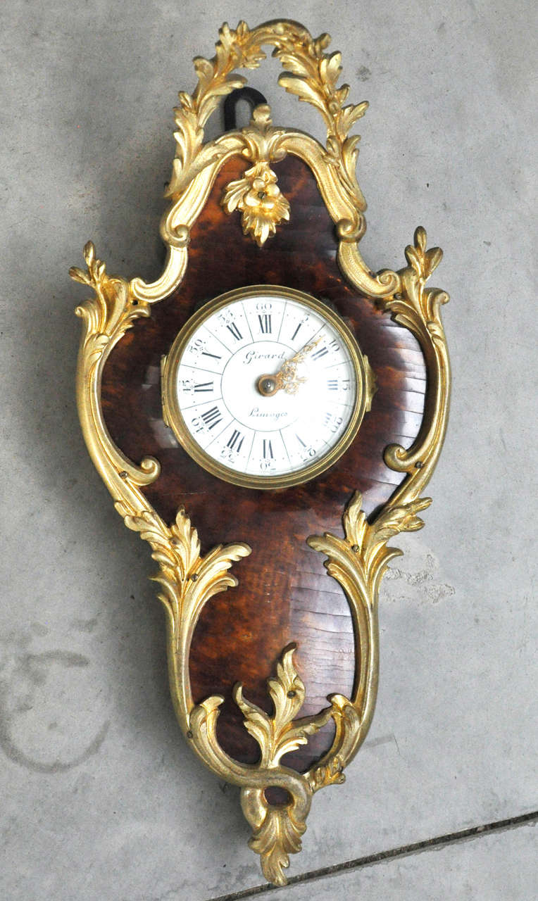 A petite cartouche shaped Cartel clock in the Louis XV style with tortoiseshell body framed with gilt bronze leafy ornamentation. Hinged convex glass door reveals white enameled dial with black Roman and Arabic numerals, pierced gilt hands, signed