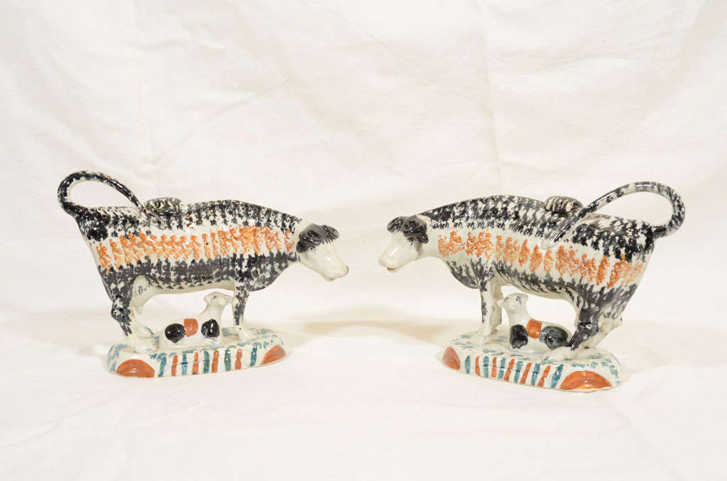 A pair of 18th century Prattware cows each with a calf resting below its mother. Sponged in horizontal bands of black and orange with vertical stripes of orange and green on the base. They were made to be creamers with a covered opening on the back