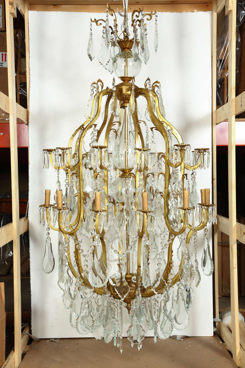 This stunning 19th Century gilt bronze chandelier is a massive 6 feet tall. It boasts scrolling candlearms extending from a glass sphered center stem and dripping with faceted drops. The twenty-four lights have tiers of obelisks surrounding each