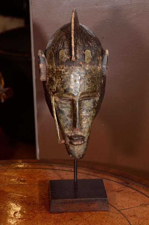 The carved wood mask mounted with a face shield of hammered,
patinated brass. The mask decorated with two brass tubes issuing tassels. The mask mounted on a stand.