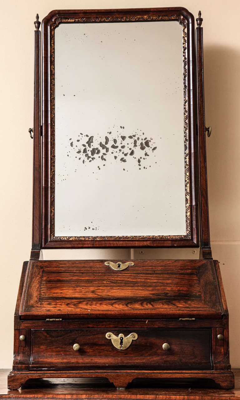 The rectangular mirror plate with cusped top corners within a conforming incised gilt surround and molded hinged wood frame set between two square spreading supports surmounted by flaming urn finials; over a slant-front case open to an arrangement