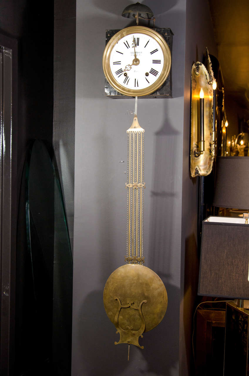 Exceptional wall clock with hand forged blackened metal frame with circular white enameled face and large brass pendulum with lyre design.  The clock also features large bell strike.