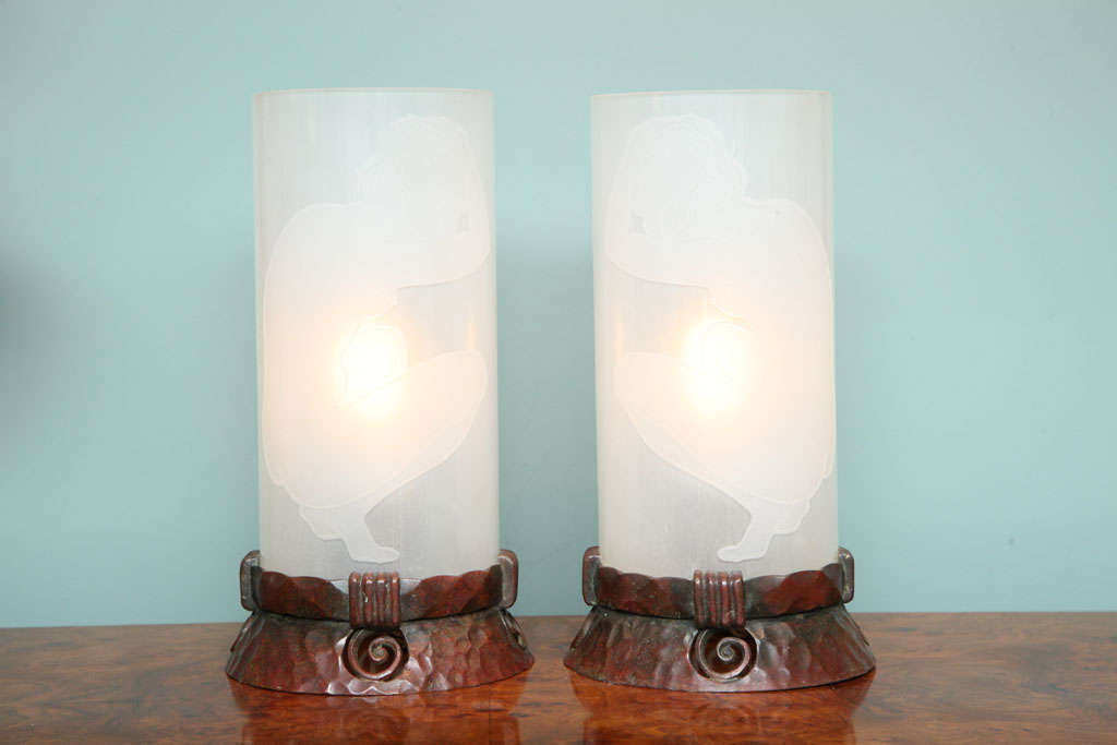 A pair of continental bronze & etched glass boudoir lamps each decorated with an acid-etched glass cylinder with a carved kneeling nude figure a top a pair of pierced bronze and hammered bases.