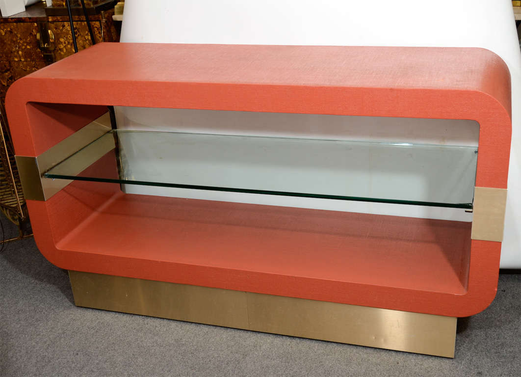 A vintage red grasscloth console table similar to works produced by Karl Springer. The piece has a single clear glass shelf and brushed brass detailing.