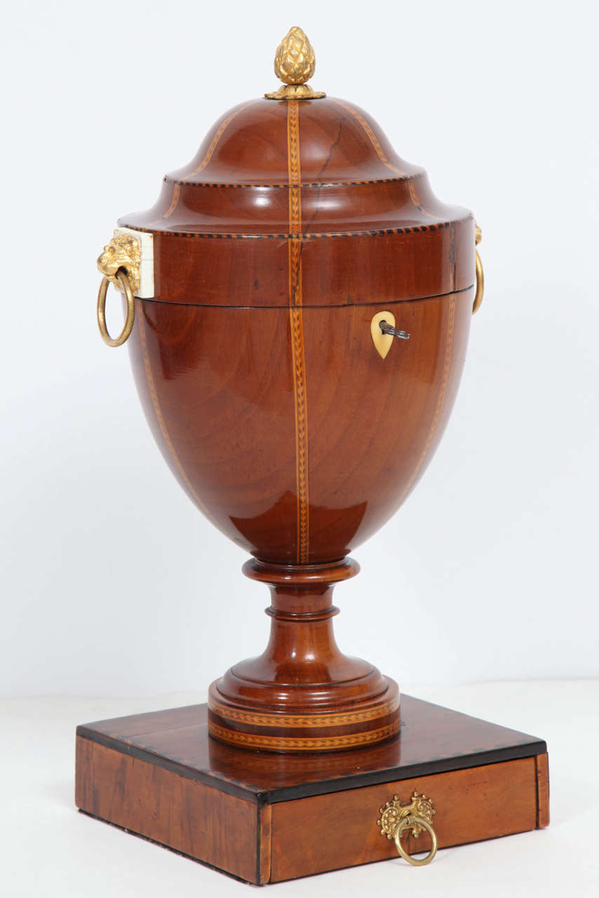 A fine Danish Empire inlaid mahogany and ormolu urn, circa 1810, of Classic form with a hinged lid surmounted with a acorn finial, the side with ring lion head mounts, raised on a square pedestal base with a drawer. Geometric fruitwood inlays though