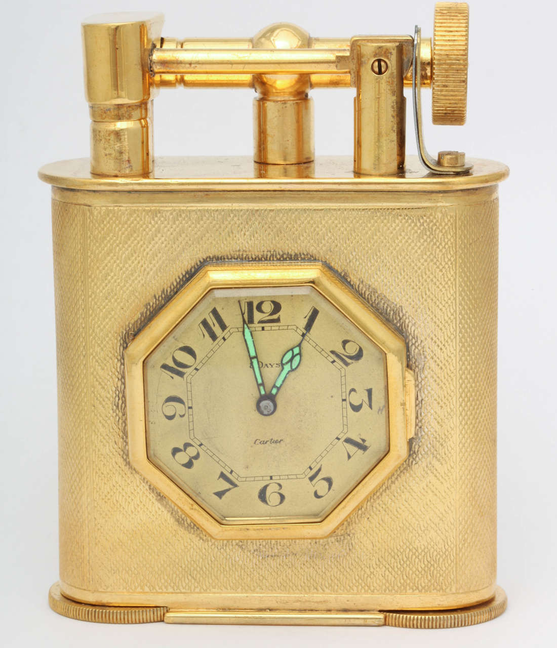 A stunning and exceptionally rare, English made, gold plated table lighter, made for and retailed by Cartier.  The lighter holds an eight day clock set in an octagonal frame in the center of the lighter's body.   The body is fully engine turned and