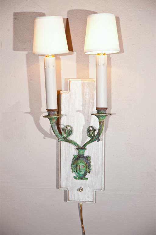 Pair of unusual French Directoire style patinated bronze two-arm wall sconces, circa 1940s; each has two candlestick arms with white linen shades, supported by bronze arms and figural-decoration, mounted on distress-painted oak plates.