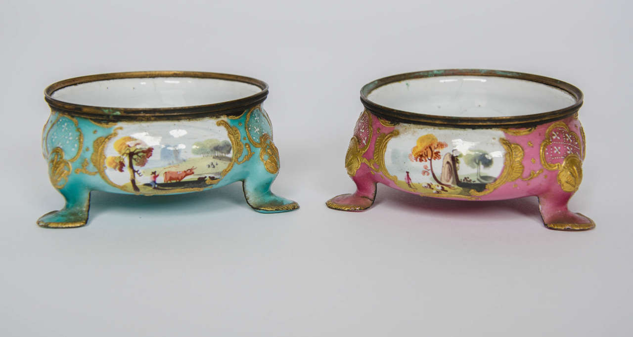 Pair of enamel Salt Cellars raised on three feet, painted with three bucolic scenes within raised gilt scroll cartouches and reserved on a pink and blue  ground, probably Bilston, South Staffordshire, circa 1780