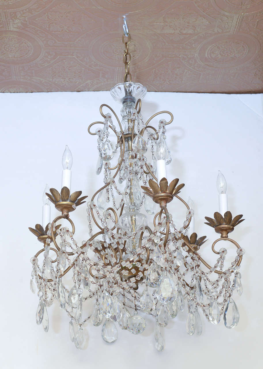 Chandelier, having a frame of gilded iron, its knopped centre-column of molded glass, six S-scroll candle arms finish with foliate bobeches, the entire piece draped with macaroni-beaded swags and faceted crystal prisms.

Stock ID: D8691